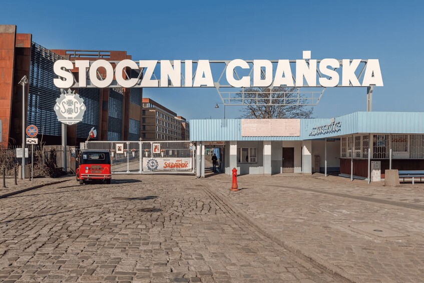 A street with a sign reading 'STOCZNIA GDANSKA' and a red bus in the foreground. 3