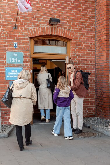 A group of people exiting a brick building with a sign reading 'Sala Konferencji Muzeum Zakładu.' 4