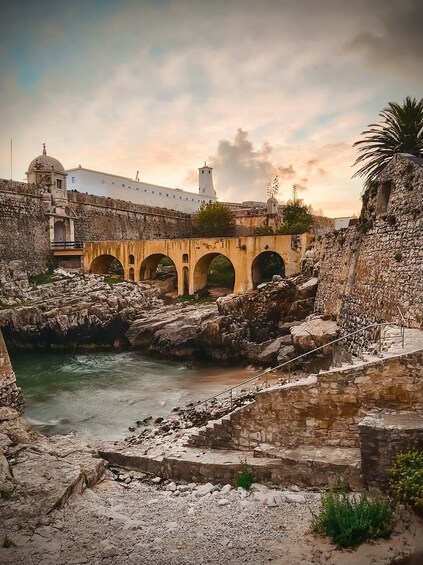 A stone bridge over a river, surrounded by rocky terrain and ancient buildings. 3