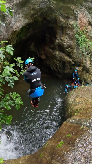 A person jumping into a river from a rocky cliff, another person in a kayak nearby, surrounded by greenery and a cave-like rock formation. 3