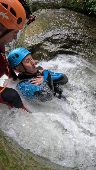 A person in a blue wetsuit and helmet, being carried by another person in a red jacket, through a turbulent water stream. 1