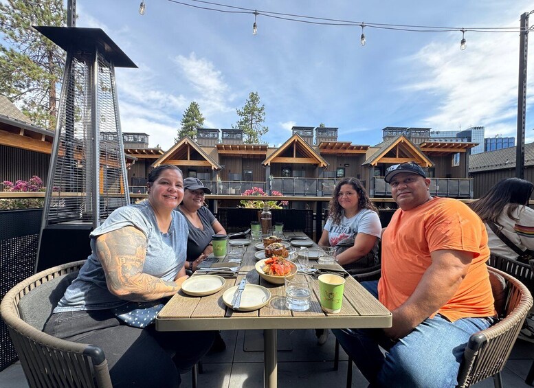 A group of people dining outdoors at a wooden table with a metal grill structure behind them. 1