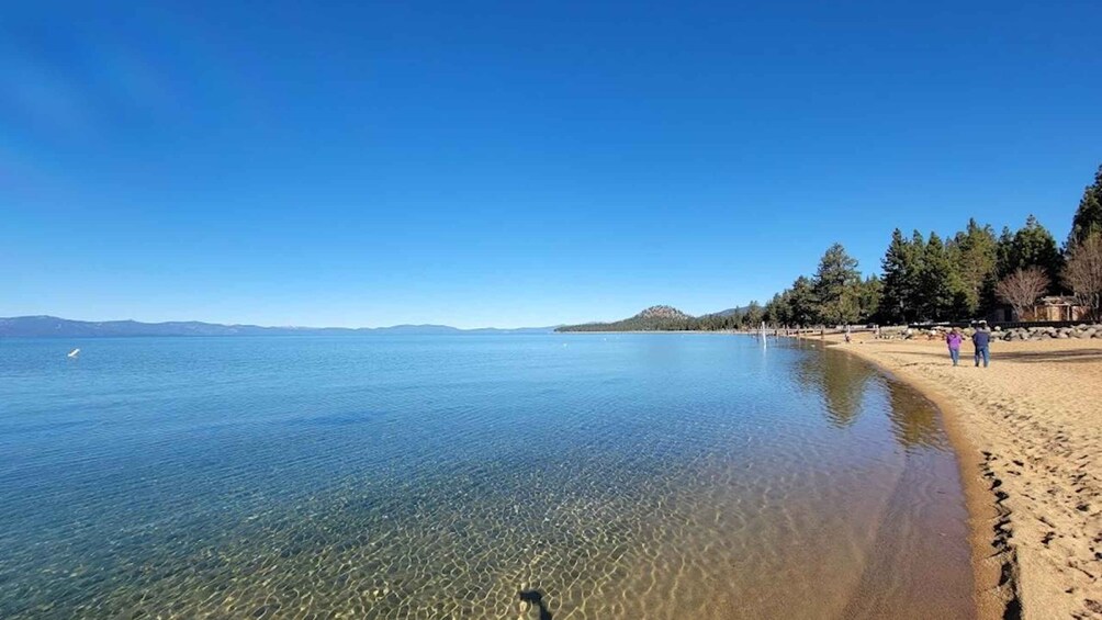 A clear lake with a sandy beach, people walking, and trees in the background. 4