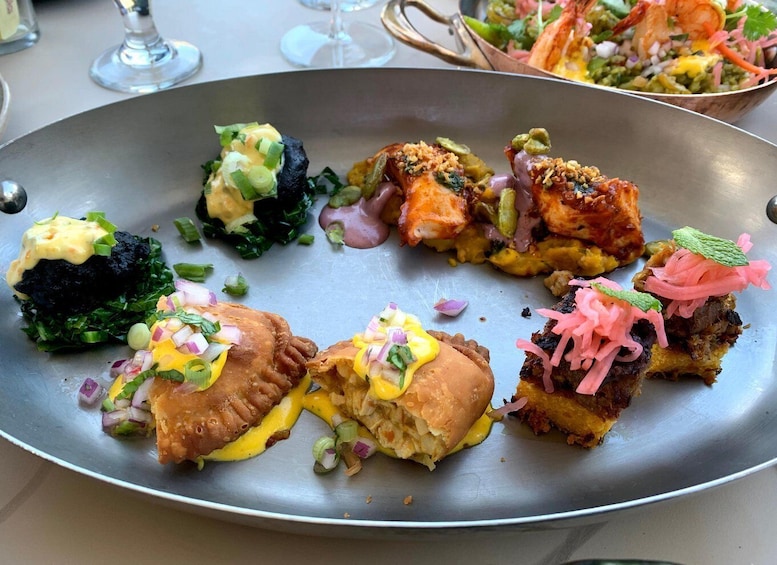 A variety of dishes on a metal plate, including a burger with yellow sauce and green onions, a fish fillet with herbs, and a salad with shrimp and vegetables. 5