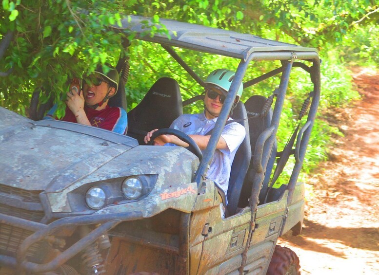 A rustic jeep with two passengers, one wearing a green cap, driving on a dirt path surrounded by greenery. 2