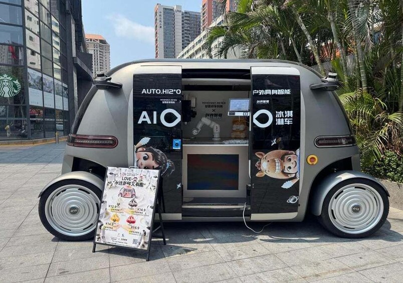 A vehicle with large wheels and a display screen, parked in an urban area with buildings and palm trees in the background. 1