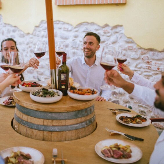 A group of people toasting with wine glasses around a wooden barrel table with various dishes. 1