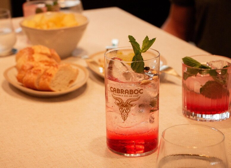 A glass of red beverage with a mint garnish, labeled 'CABRABOC,' on a table with a bowl of food and a plate of bread. 2