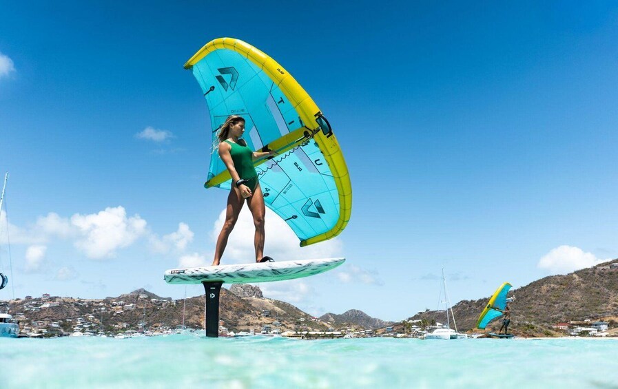 A person windsurfing on a clear day with a blue and yellow sail, standing on a white surfboard, with a coastal town and hills in the background. 2