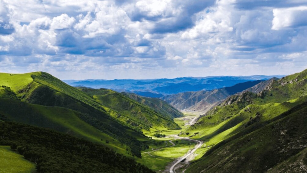 A valley with a winding road, green hills, and distant mountains under a cloudy sky. 3