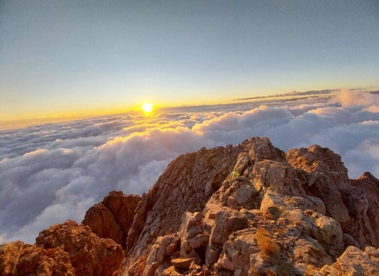 A mountain peak with clouds below and the sun setting in the background. 2