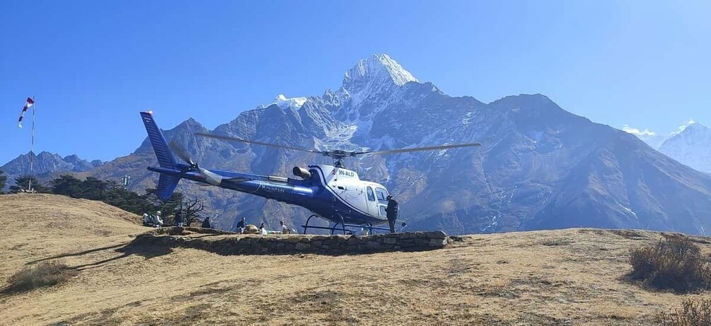 A blue and white helicopter parked on a grassy hill with a mountainous backdrop. 1