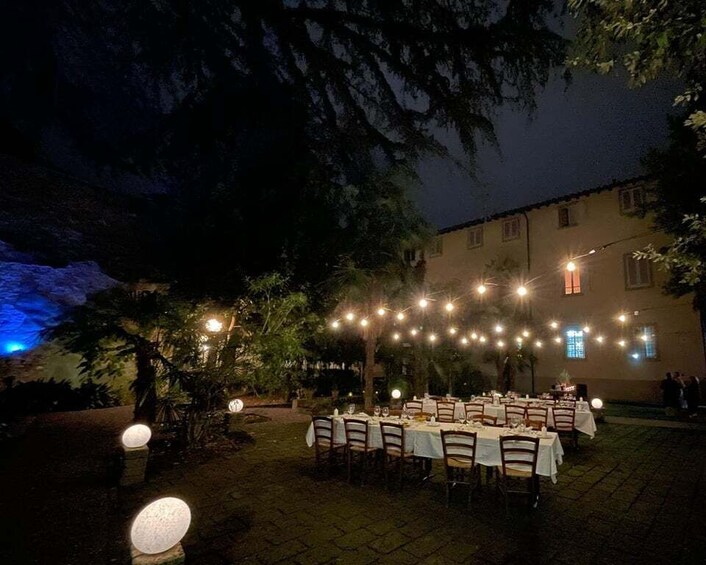 An outdoor dining area with tables and chairs, string lights, and a building in the background. 1