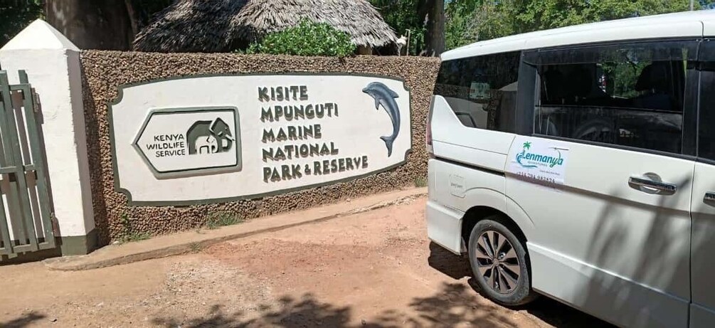 A sign for Kisite Marine National Park & Reserve with a dolphin graphic, a white vehicle parked nearby, and a thatched-roof structure in the background. 4