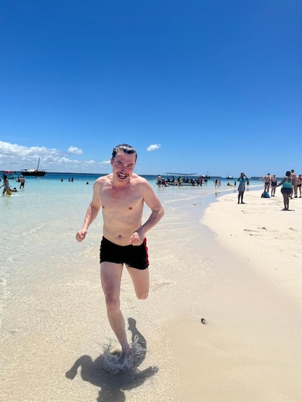 A man running on a sandy beach with clear blue water and boats in the distance. 3