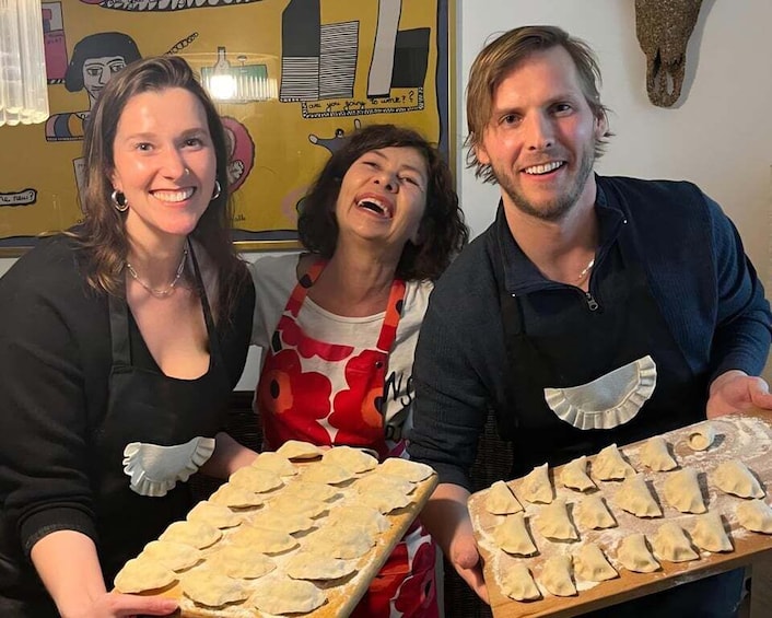 Three people holding trays of food, possibly pastries, in a kitchen setting with a wall mural in the background. 1