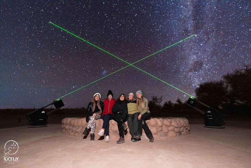 A group of people posing under a night sky with visible stars and the Milky Way, with laser beams crossing the sky. 4