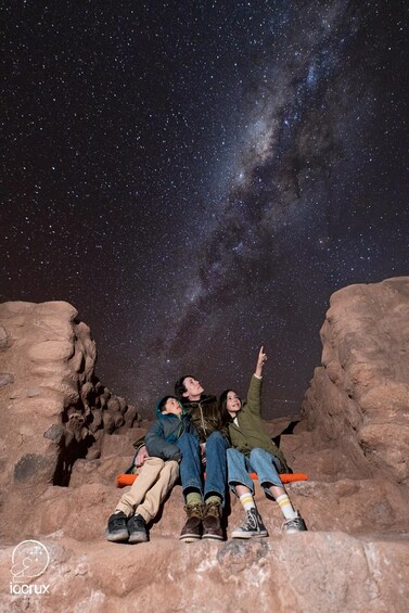 Three people sitting on a rocky outcrop under a starry night sky. 5