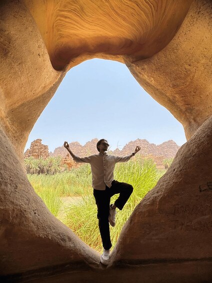 A person standing inside a natural rock formation with arms outstretched, surrounded by desert vegetation and mountains. 2