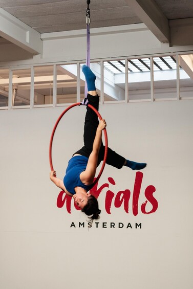 A person performing aerial acrobatics with a hoop in a studio with a sign reading 'Aerials Amsterdam'. 5