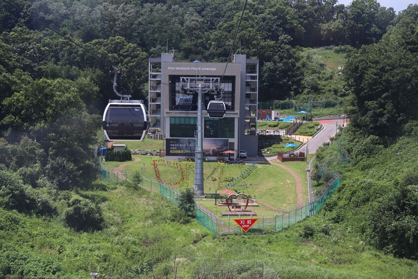 A cable car station named 'PAJU INJINGAK PEACE GONDOLA' with a cable car in motion, surrounded by lush greenery and a winding road. 2
