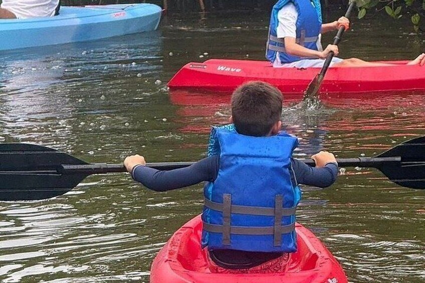 A child in a red kayak wearing a blue life jacket, paddling on calm water. 2