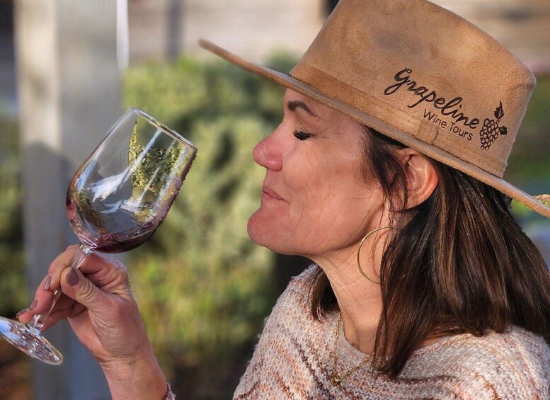 Henkilö, jolla on kädessään viinilasi, jossa on viinitarhan logo, ja hänellä on päässään hattu, jossa lukee teksti »Grapeline Wine Tours». 3