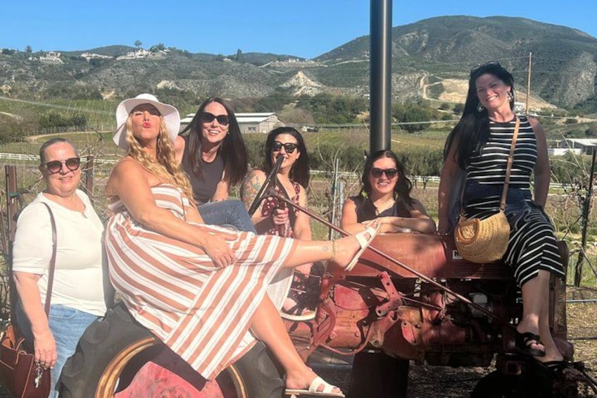A group of people sitting on a vintage tractor in a vineyard with hills in the background. 5