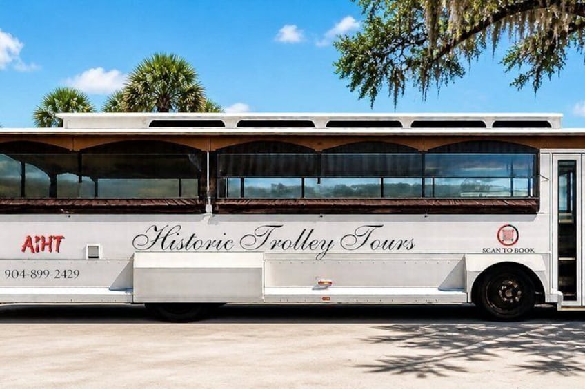 A white bus with 'Historic Trolley Tours' written on the side, parked on a concrete surface with trees in the background. 1