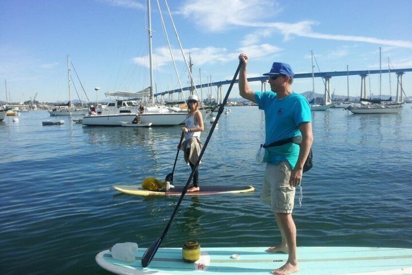 Two people paddleboarding on calm water with a bridge in the background. 3