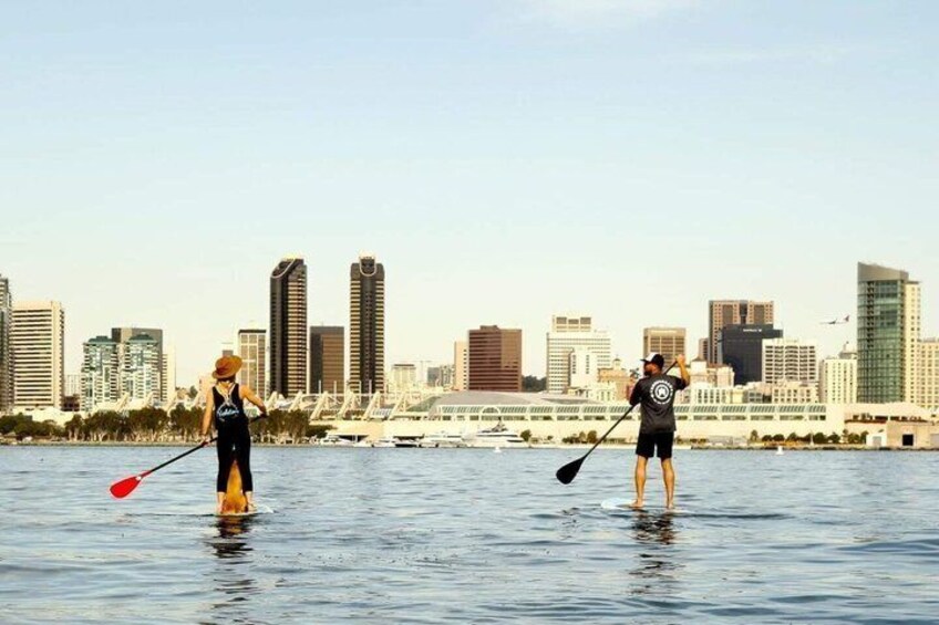 Two people paddleboarding on a body of water with a city skyline in the background. 1