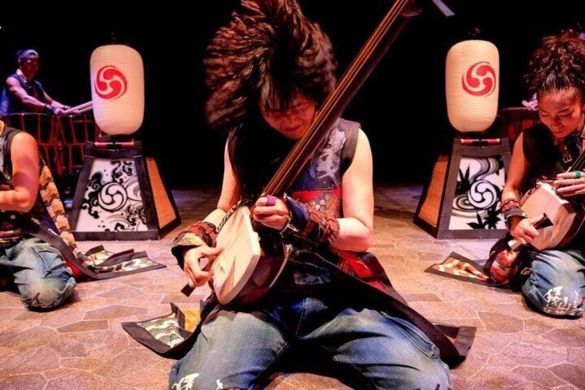 A person playing a stringed instrument, seated on the floor with a patterned backdrop. 5