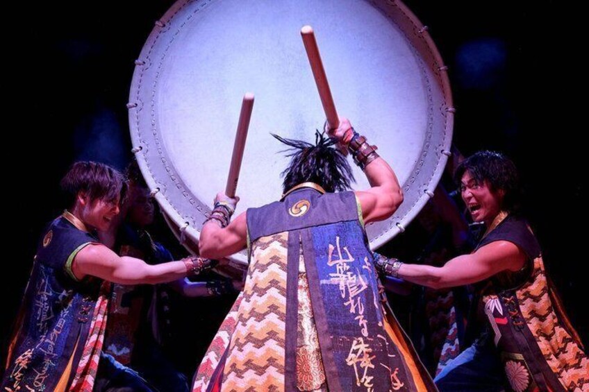 A person in traditional attire with a large drum and two sticks. 4