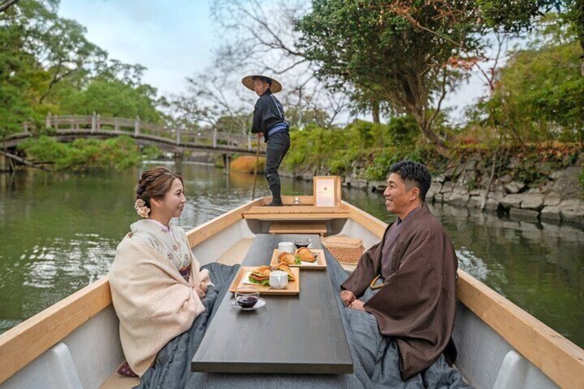 A traditional wooden boat with two people dining, a person standing on the dock, and a bridge in the background. 4
