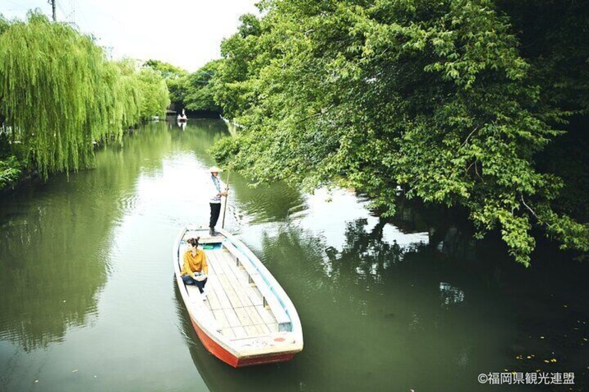 A narrow waterway with a small boat, a person standing at the stern, and dense greenery on both sides. 1