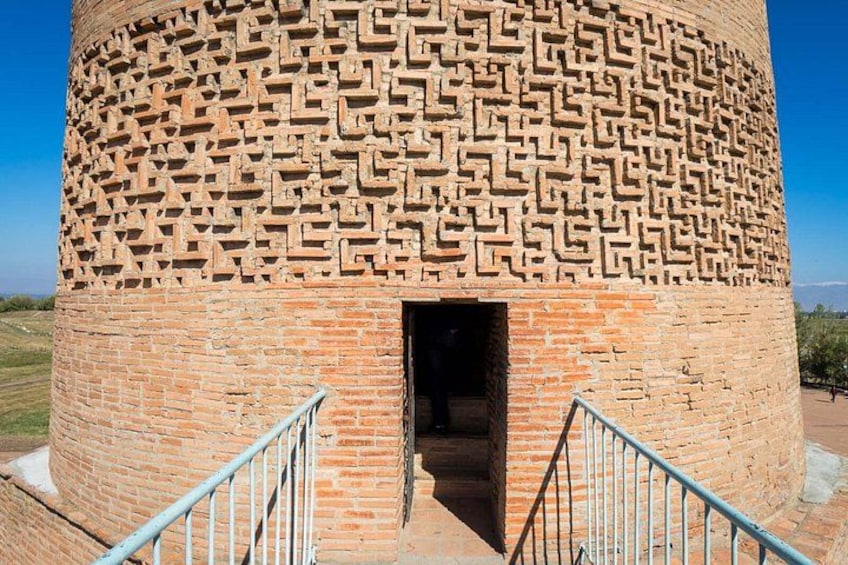 A cylindrical brick structure with a maze-like pattern on its exterior, a metal staircase leading up to an entrance, and a clear blue sky in the background. 4