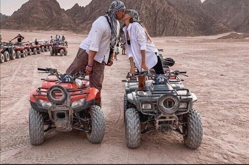 Two people on ATVs in a desert landscape with rocky terrain and distant mountains. 1