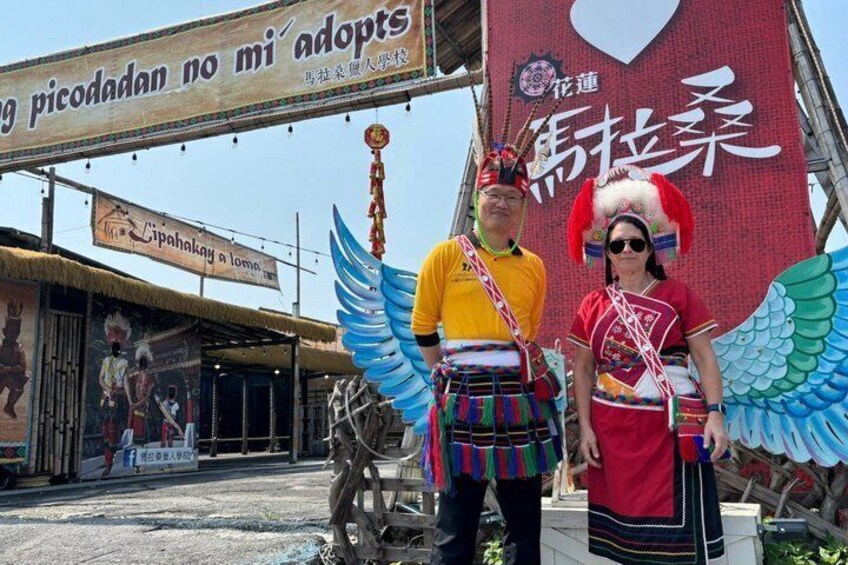 Two individuals in traditional attire with wings, standing in front of a sign with text in a foreign language. 1