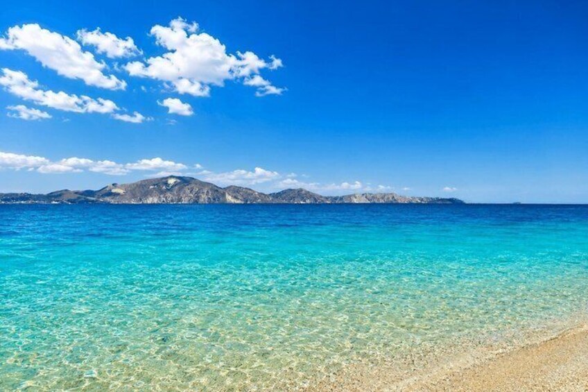 A clear turquoise sea with a sandy beach and distant mountains under a blue sky with clouds. 4