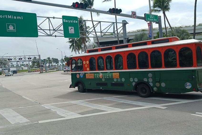 A green and orange Miami Trolley at an intersection with overhead signs for U.S. 1 and Bayside Blvd. 5
