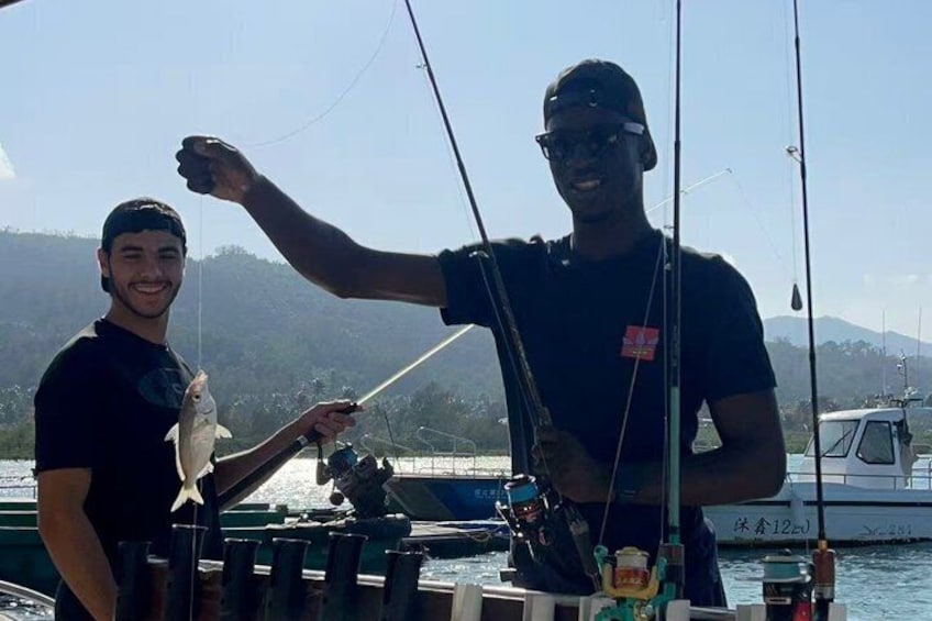 Two people on a boat holding fish, with a dock and a boat in the background. 1