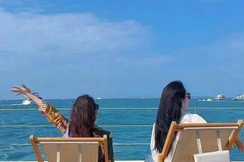 Two people sitting on lounge chairs facing the sea, with boats in the background. 5