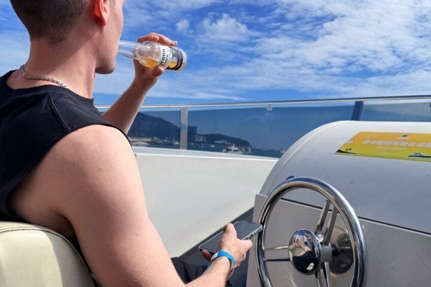 A person in a black sleeveless top, holding a bottle, and steering a vehicle with a steering wheel and a yellow sign on the dashboard. 3