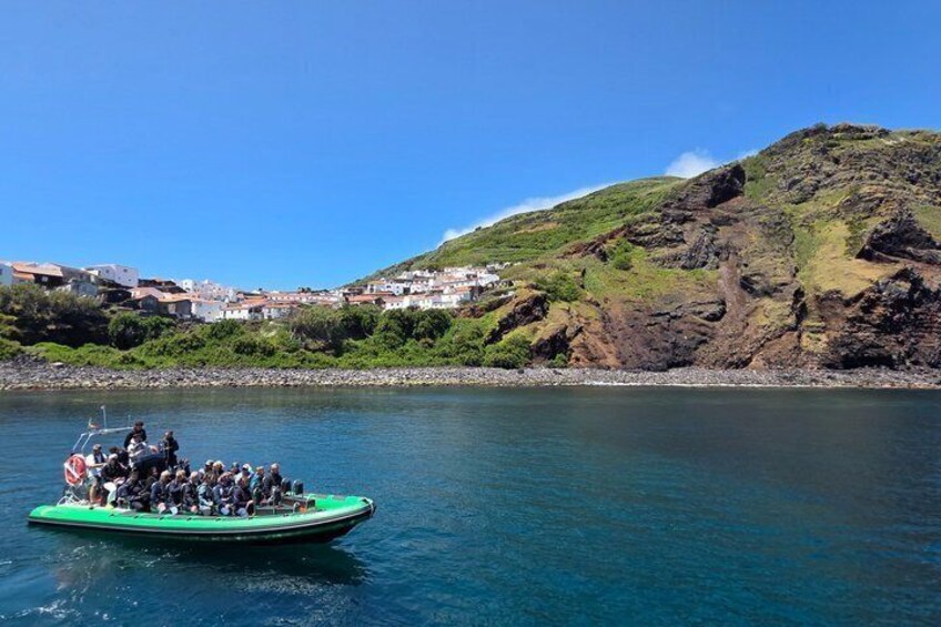 A green boat with passengers on a calm body of water, surrounded by a coastal landscape with buildings and hills. 4