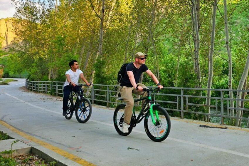 Two people riding bicycles on a paved path surrounded by trees. 1