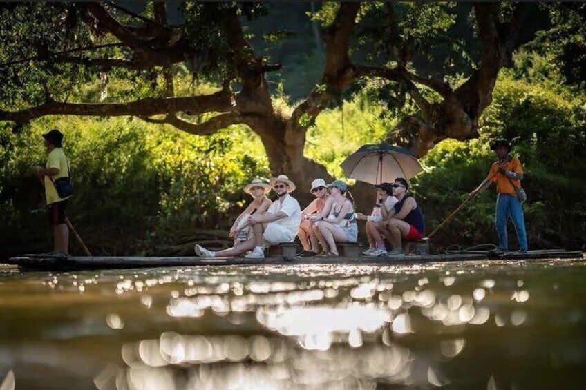 People sitting by a calm body of water, some holding umbrellas, with trees and a person standing in the background. 4