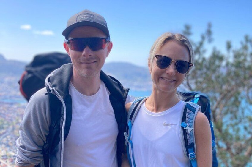 Two people with backpacks, one wearing a cap, standing in a mountainous area with trees and a distant cityscape. 2