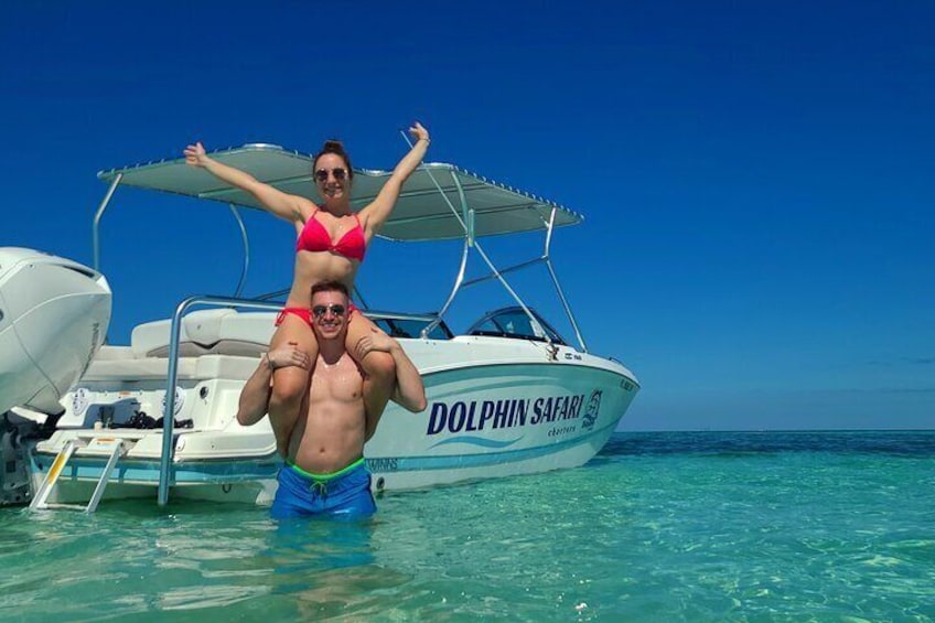 A man carrying a woman on his shoulders in front of a boat named 'Dolphin Safari.' 1