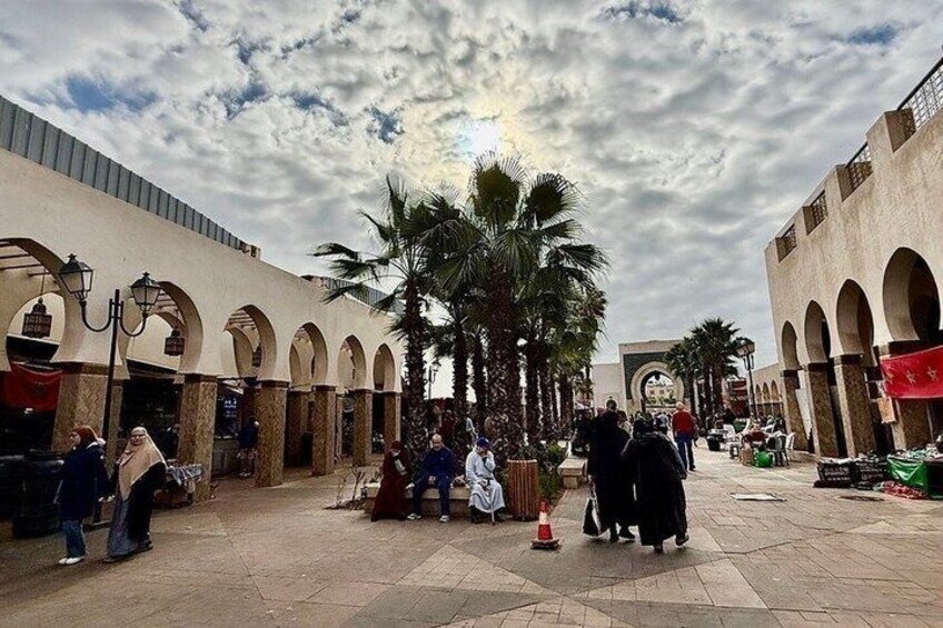A bustling market street with palm trees, arches, and people shopping. 4