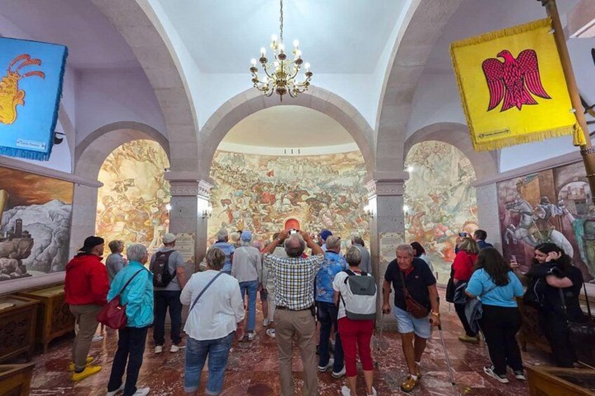 A group of people inside a museum with arches, a chandelier, and a large mural. 3